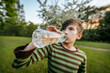 © Emvats - young boy drinking water from a plastic bottle, wearing a striped shirt, with a blurred green outdoor background.