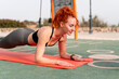 © Andrii - Happy fit strong woman with red hair doing plank exercise on mat. Side view of female athlete in sportswear doing fitness exercise outdoors on sunny summer day.
