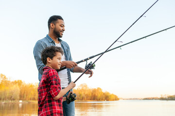 african american man and child sitting on a wooden pier holding fishing rods on the river, dad teach