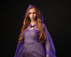 Wall Mural - close up portrait of beautiful female model with long red hair, wearing a purple fairy  fantasy cloak with hood and gown. Elegant gestural hand poses as if casting magic spell. Isolated on dark black 