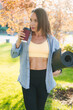 © Strelciuc - Beautiful caucasian adult woman with brown hair standing in summer park holding yoga mat drinking a cup of takeaway tea or coffee looking away.