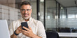 © Stock 4 You - Banner with middle-age Hispanic business manager ceo using cell phone mobile app. Smiling Latin or Indian mature man businessman holding smartphone in office, working as trader on pc, with copy space
