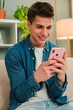 © Jose Calsina - Vertical. Young guy smiling using his smartphone to browse on internet at home living room sitting on a sofa. Male betting online, texting messages or sharing on the social media with a cellphone app