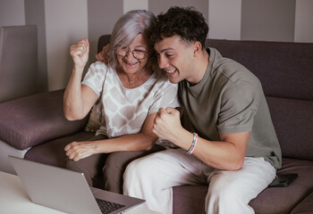 Cheerful couple of boy and elderly grandmother watching summer olympic games together on laptop rejoicing for victory sitting on comfortable sofa in living room