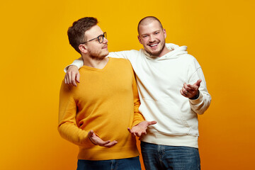 Two cheerful young men embracing each other, One guy looking with uncertainty have doubtful facial expressions, second male giving solution. Isolated over yellow background