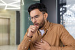 © Tetiana - Young man coughing in modern office environment, hand on chest