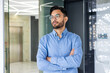 © Tetiana - Confident businessman in blue shirt standing with crossed arms in modern office environment