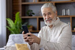 © Tetiana - Portrait of senior man, grandfather at home, sitting on sofa and using phone, smiling and looking at camera