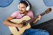 © Marcos - Young latin man playing acoustic guitar at home in living room using headphones in Mexico Latin America