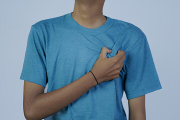 close up portrait of young Asian man in blue shirt moaning in pain while holding his chest, heart disease, isolated in gray background.