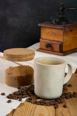 Still life of a cup of coffe sitting in the foreground on a heap of roasted coffee beans and a jar with brown sugar. In the background there is an old coffee grinder. They all sit on a white cotton cl