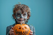 © AISTEL - photo of a happy boy with zombie face painting, dressed in gray suit and holding halloween pumpkin on blue background