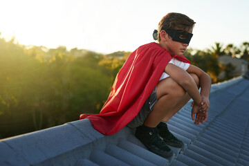 Serious, young boy and playing superhero on roof with costume and mask for fantasy adventure at home. Male person or child and hero game for protection, safety and justice of neighbourhood in summer