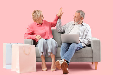 Poster - Mature couple with laptop and shopping bags giving each other high-five on sofa against pink background