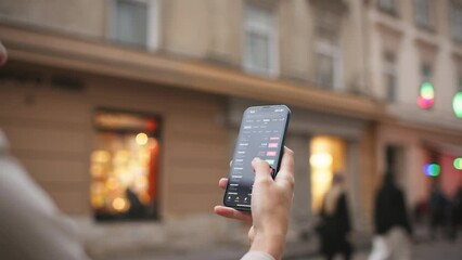 Side view of woman holding smartphone while standing in middle of street in city. Looking at screen while comparing price changes on stock market. Concept of cryptocurrency. Online trading.