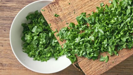 Poster - Chef puts fresh green chopped parsley leaves in bowl from wooden cutting board to prepare healthy food, top view. Food close up