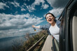 © qunica.com - Excited young woman screaming with joy and holding a beverage, leaning out of a car window against a backdrop of clouds and mountains during a road trip.