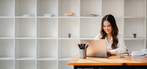 Wall Mural - A woman is sitting at a desk with a laptop and a pencil holder. She is smiling and she is enjoying her work. The room is filled with shelves, and there are several books scattered around