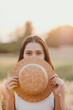 © Satori Studio - A woman is holding a straw hat and is hiding her face behind it. The hat is brown and has a round shape. The scene is set in a field with grass and trees in the background