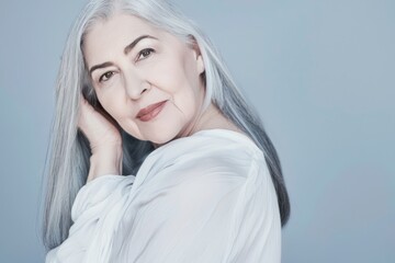 Elder female model, left hand under long gray hair, smiling senior woman face looking at camera, in white blouse, on blue background. Happiness and attractiveness in age.