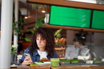 Woman with curly hair seats at vegetarian cafe or restaurant with green smoothie and plant meal on table, looking smartphone. Venues VEGAN sign displayed in background, healthy dining environment