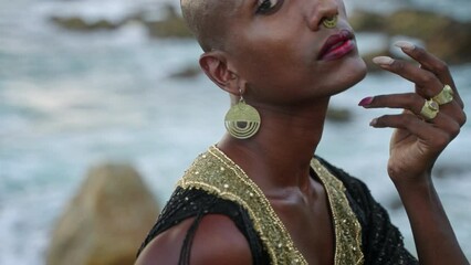 Wall Mural - Gender fluid person of color in luxury dress, jewelry poses on stormy ocean shore. Bossy extravagant fashion model in posh gown, shows hand with golden rings, gems, bracelet, looks at camera.