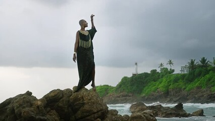 Wall Mural - Divine non-binary black person in boutique dress brass jewelry poses on rocky hill top above dramatic ocean cloudy skyline. Queer lgbtq fashion model in posh outfit fluttering in air. Pantheon concept