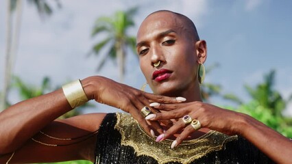 Wall Mural - Gay bipoc man in luxury dress boutique brass jewelry, makeup poses looking on camera with moody face. Homosexual ethnic elegant person in posh gown wears nose-ring piercing, earrings. Palm trees.