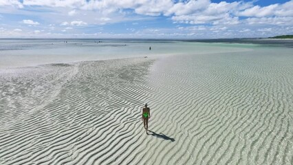 Wall Mural - Aerial view of walking young woman on the sandbank in ocean, white sand, blue sea during low tide at sunny summer day in Zanzibar island. Top view of girl, sand spit, bay, clear water, sky with clouds