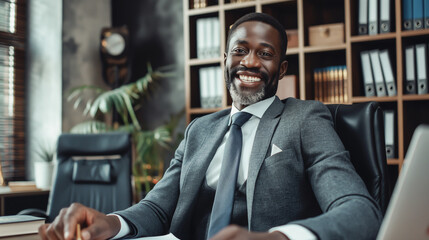 A young successful African American businessman in a business suit sits at a desk in his office against the backdrop of bookshelves and smiles softly, demonstrating professionalism and experience.