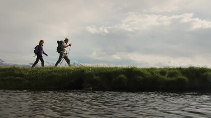 Fototapete - Silhouettes of two adult hikers with backpacks walks at riverside