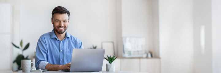 Poster - Smiling Young Businessman Sitting At Desk With Laptop In Modern Office, Looking At Camera, Free Space