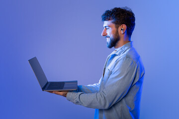 Poster - A man with a beard is standing while holding a laptop in his hands. He appears to be looking at the screen intently, possibly working or browsing the internet.