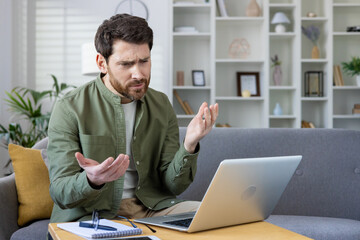 Wall Mural - Frustrated man working from home on laptop with confused expression. Young professional experiencing technical difficulties while working remotely in a modern living room setup.