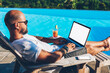© BullRun - Businessman enjoying hot tour during summer resort lying on sunbed with cocktail near pool while earning money doing distance job at netbook with blank copy space screen for your advertising content