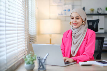 a woman wearing a pink jacket and a head scarf is sitting at a desk with a laptop. she is smiling an