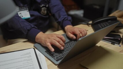 Hispanic detective in uniform working on laptop in a cluttered police station office.