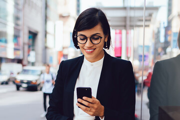 Wall Mural - Cheerful female manager getting to work by foot drinking morning coffee to go while banking online on telephone.Successful entrepreneur in elegant wear reading incoming message on smartphone