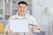 © Liubomir - Happy man showcasing a blank whiteboard in a comfortable living room setting while holding a pen.