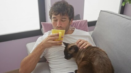 Poster - A relaxed man enjoys coffee while petting a siamese cat on a couch indoors