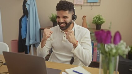 Sticker - Hispanic man with a beard celebrates success while wearing headphones and working on a laptop at home.