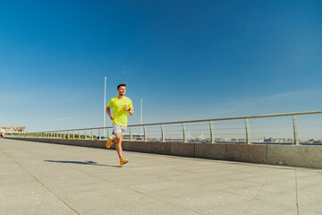 A man in a yellow shirt and shorts jogs along a sunny waterfront promenade, with clear blue skies and a city skyline in the distance.