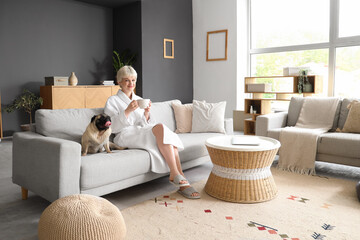 Sticker - Young woman in bathrobe with coffee cup and pug dog sitting on sofa at home