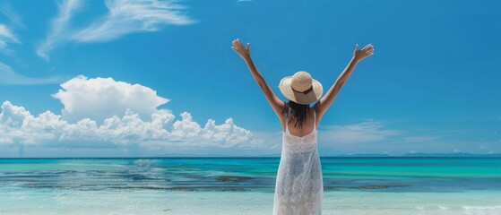 Wall Mural - A woman stands on the beach with her arms raised, embracing the serene beauty of the ocean under a bright blue sky filled with fluffy clouds. Perfect for travel and vacation themes.