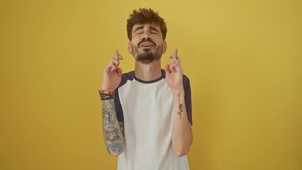 Sticker - Young hispanic man standing with fingers crossed for luck, eyes closed with hope, making a superstitious gesture on an isolated yellow background