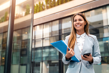 Young adult smiling professional business woman wearing suit holding smartphone and folder with files while walking in urban city environment.