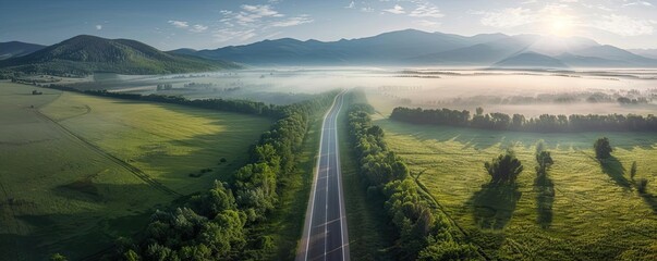 Poster - Aerial view of a straight country road through lush green fields and mountains in the background on a misty morning.