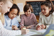 © AnnaStills - Group of ethnically diverse kids sitting at table in classroom doing exercise together during English lesson