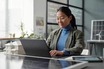 Aufkleber - Smiling Asian female employee looking at laptop screen and typing while sitting by workplace with house model and solar panel