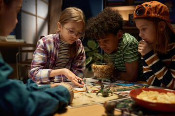 Group of serious intercultural children bending over table while one of them throwing dice on game board with cards and figurines